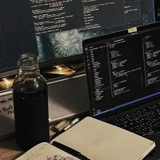 Gece bir masa: dizüstü bilgisayar, kod dolu bir monitör ve açık bir defter